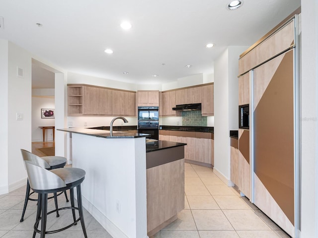 kitchen featuring double oven, light tile patterned floors, light brown cabinets, paneled built in fridge, and an island with sink