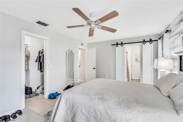 tiled bedroom featuring a walk in closet, a barn door, ceiling fan, a closet, and ensuite bathroom