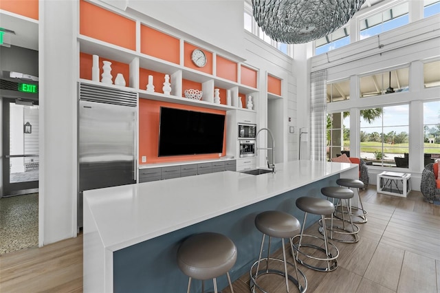 kitchen featuring a towering ceiling, built in refrigerator, light wood-type flooring, sink, and a breakfast bar