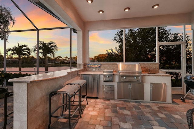 patio terrace at dusk featuring a water view, an outdoor kitchen, a grill, and a lanai