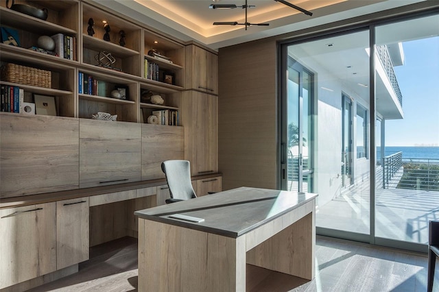 office area with ceiling fan, light hardwood / wood-style floors, a raised ceiling, built in desk, and a water view