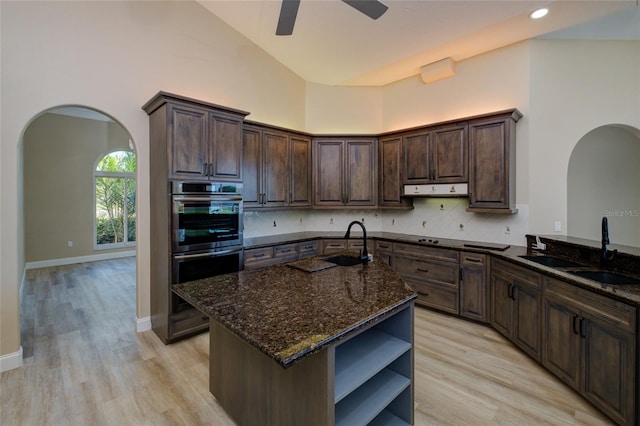 kitchen with backsplash, an island with sink, dark stone countertops, double oven, and ceiling fan