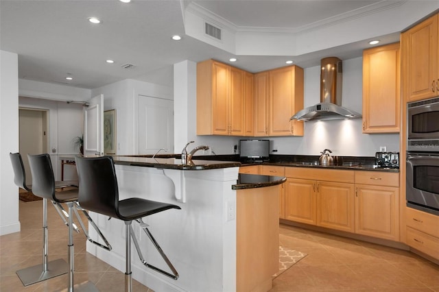 kitchen featuring dark stone countertops, a breakfast bar area, wall chimney exhaust hood, and light brown cabinets