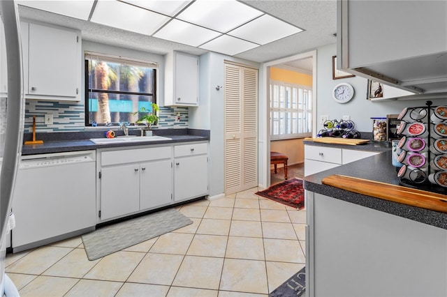 kitchen featuring light tile patterned floors, dark countertops, white dishwasher, white cabinetry, and a sink