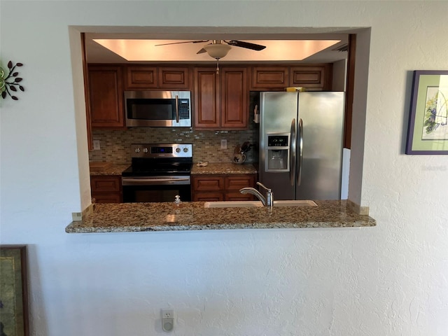 kitchen with appliances with stainless steel finishes, light stone counters, backsplash, ceiling fan, and a raised ceiling