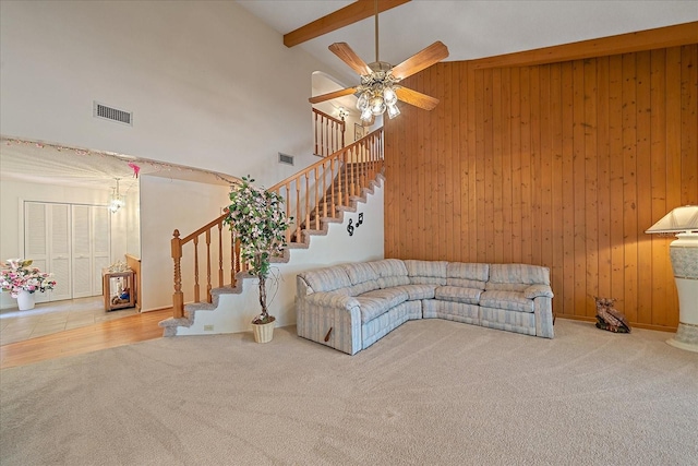 carpeted living room featuring ceiling fan, a towering ceiling, wooden walls, and beam ceiling