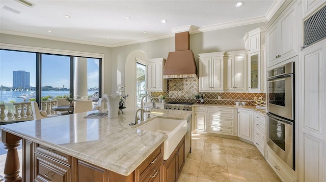 kitchen with appliances with stainless steel finishes, a large island with sink, wall chimney exhaust hood, and ornamental molding