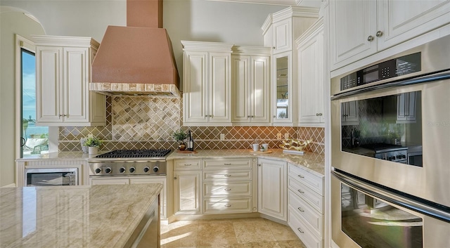 kitchen with light stone counters, stainless steel appliances, custom range hood, and tasteful backsplash