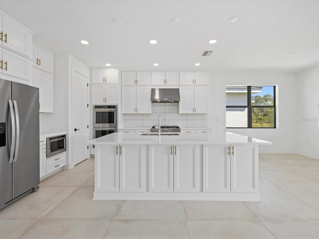 kitchen featuring a center island with sink, backsplash, wall chimney range hood, white cabinetry, and stainless steel appliances