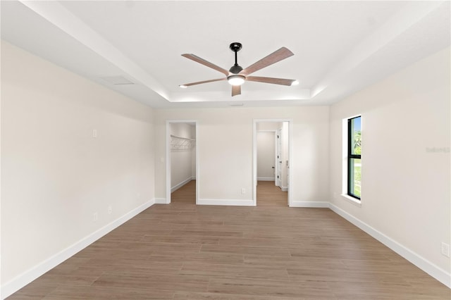 unfurnished bedroom featuring baseboards, light wood finished floors, a walk in closet, a raised ceiling, and a ceiling fan