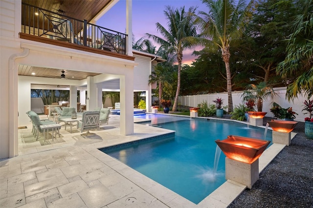 pool at dusk with pool water feature, ceiling fan, an outdoor hangout area, and a patio