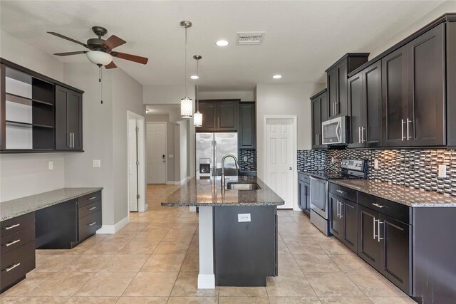 kitchen featuring a kitchen island with sink, dark stone counters, hanging light fixtures, sink, and appliances with stainless steel finishes