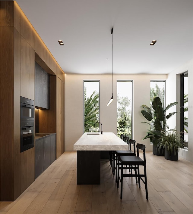 interior space featuring hanging light fixtures, black double oven, light wood-type flooring, a center island with sink, and a breakfast bar area