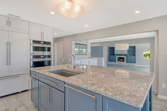 kitchen featuring light stone countertops, decorative light fixtures, a center island with sink, stainless steel double oven, and sink