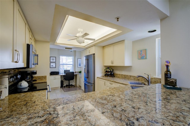 kitchen with light stone counters, stainless steel appliances, ceiling fan, sink, and a tray ceiling