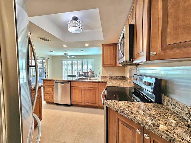 kitchen with sink, ceiling fan, light stone counters, appliances with stainless steel finishes, and a tray ceiling