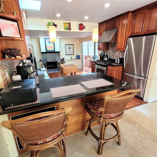 kitchen with appliances with stainless steel finishes, wall chimney exhaust hood, a tile fireplace, and pendant lighting