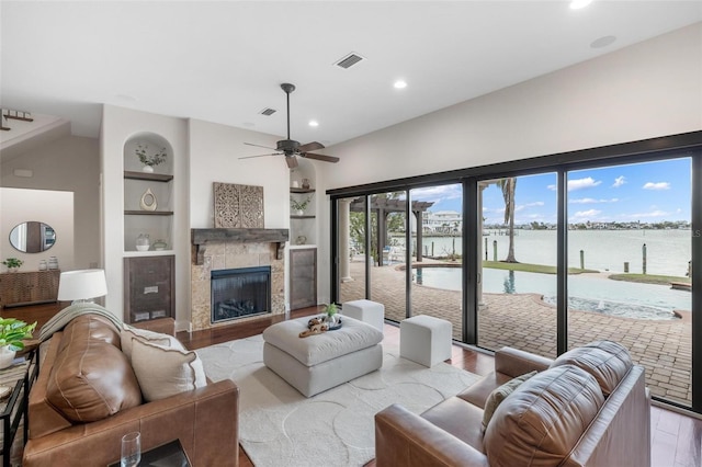 living room featuring built in shelves, ceiling fan, a stone fireplace, light hardwood / wood-style floors, and a water view