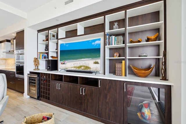 bar with light wood-type flooring, dark brown cabinetry, stainless steel double oven, and beverage cooler