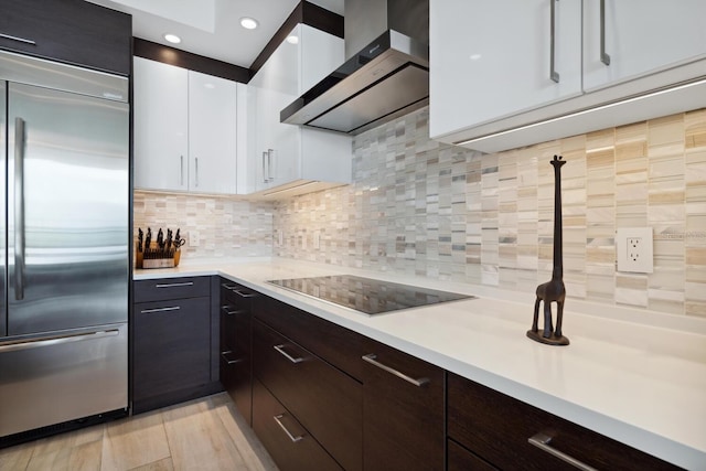 kitchen featuring wall chimney range hood, stainless steel built in refrigerator, backsplash, black electric cooktop, and white cabinets