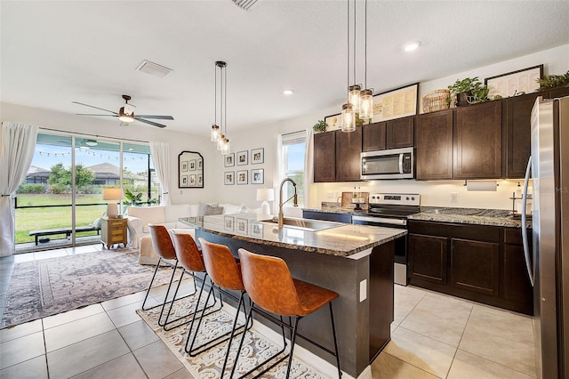 kitchen featuring ceiling fan, a center island with sink, stainless steel appliances, light tile flooring, and light stone countertops