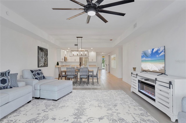 living area with a tray ceiling, visible vents, light wood-style flooring, and ceiling fan with notable chandelier