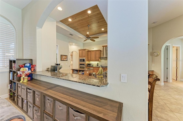 kitchen with appliances with stainless steel finishes, wall chimney exhaust hood, dark stone countertops, wooden ceiling, and light tile patterned floors