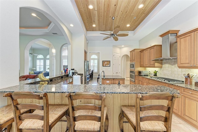 kitchen featuring appliances with stainless steel finishes, wall chimney range hood, kitchen peninsula, ceiling fan, and stone counters