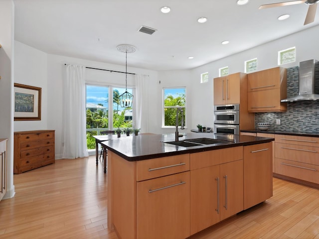 kitchen featuring hanging light fixtures, a center island with sink, wall chimney range hood, stainless steel double oven, and sink