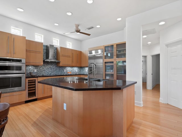 kitchen with appliances with stainless steel finishes, wall chimney range hood, a kitchen island with sink, and wine cooler