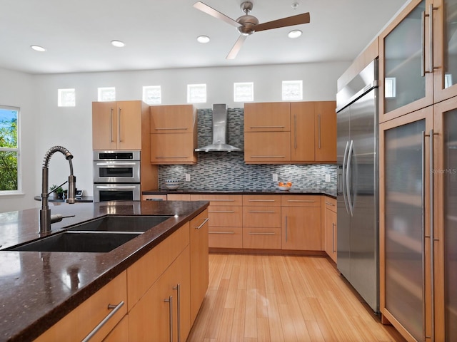 kitchen featuring wall chimney exhaust hood, dark stone counters, appliances with stainless steel finishes, ceiling fan, and sink