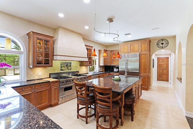 kitchen with a sink, high quality appliances, brown cabinetry, dark stone countertops, and custom range hood