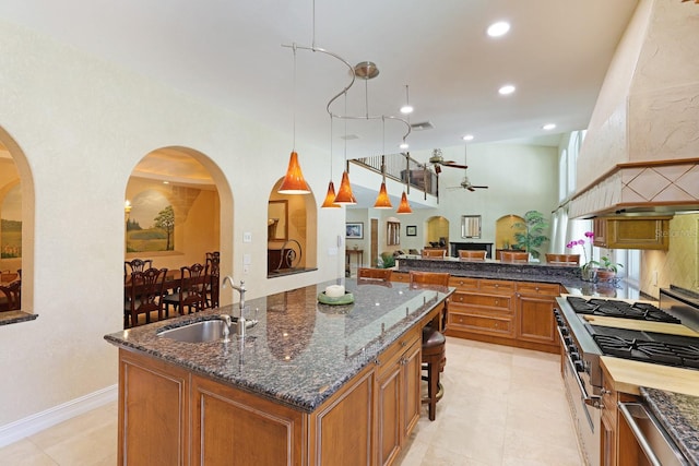 kitchen featuring a sink, high end stainless steel range oven, brown cabinetry, dark stone countertops, and a center island with sink