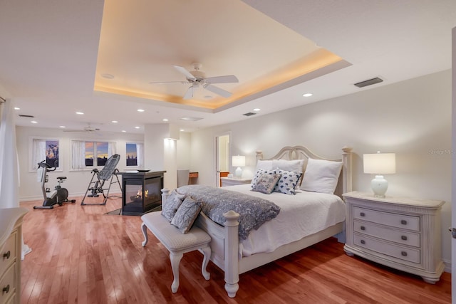 bedroom featuring hardwood / wood-style floors, ceiling fan, a multi sided fireplace, and a tray ceiling