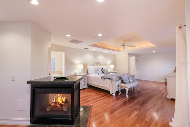 bedroom with hardwood / wood-style floors, ceiling fan, a tray ceiling, and a multi sided fireplace