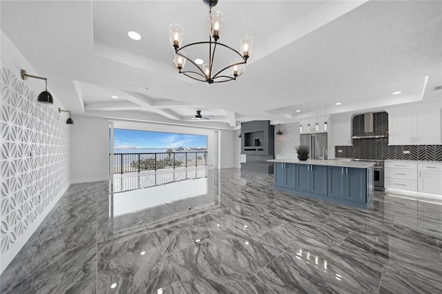 interior space with wall chimney exhaust hood, white cabinetry, a raised ceiling, hanging light fixtures, and a kitchen island with sink