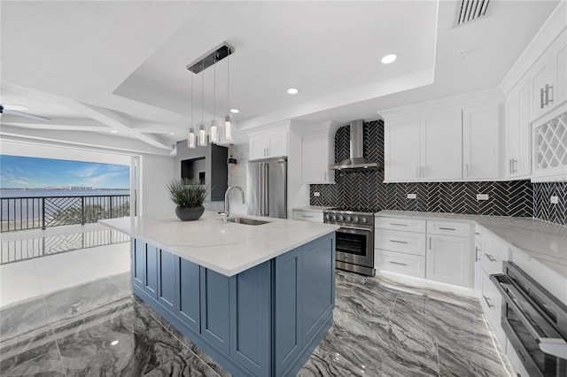 kitchen with appliances with stainless steel finishes, a center island with sink, light tile flooring, wall chimney range hood, and white cabinetry