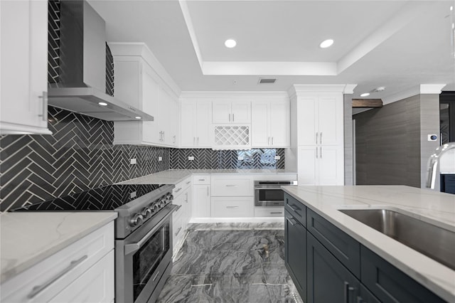 kitchen with wall chimney exhaust hood, white cabinets, appliances with stainless steel finishes, a raised ceiling, and dark tile flooring