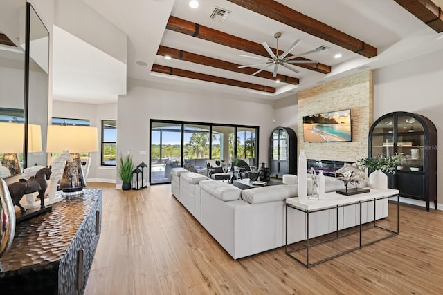 living room featuring a stone fireplace, a towering ceiling, ceiling fan, beam ceiling, and light wood-type flooring