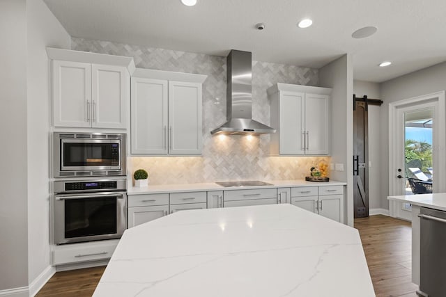 kitchen featuring white cabinetry, appliances with stainless steel finishes, a barn door, dark wood-type flooring, and wall chimney exhaust hood