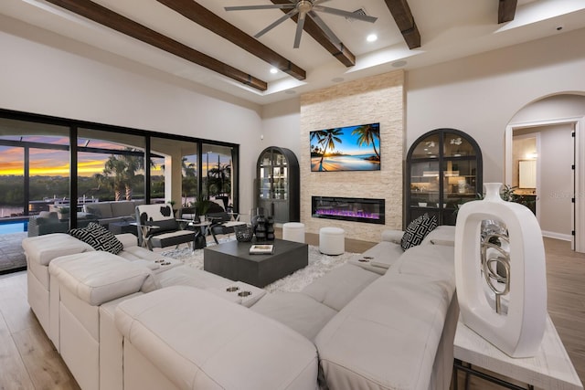 living room with a stone fireplace, beamed ceiling, ceiling fan, and light hardwood / wood-style floors