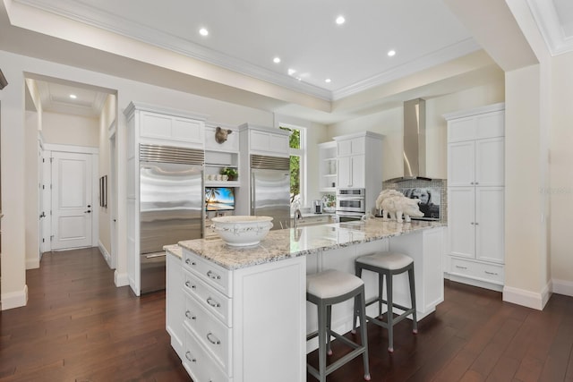 kitchen featuring white cabinetry, light stone countertops, stainless steel appliances, wall chimney range hood, and a spacious island