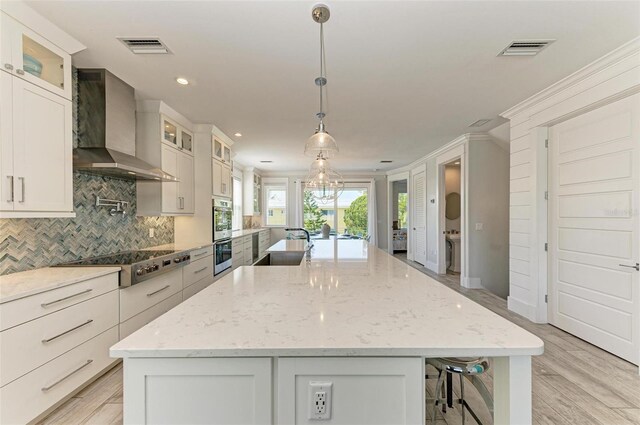 kitchen with wall chimney exhaust hood, pendant lighting, white cabinets, appliances with stainless steel finishes, and an island with sink