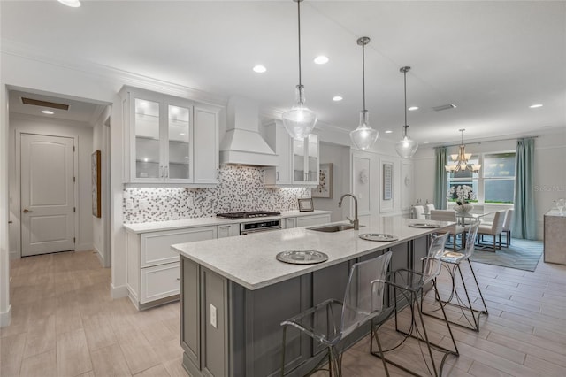 kitchen with premium range hood, decorative light fixtures, sink, tasteful backsplash, and an island with sink