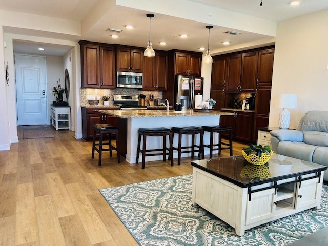 kitchen featuring backsplash, stainless steel appliances, a center island with sink, light hardwood / wood-style floors, and hanging light fixtures