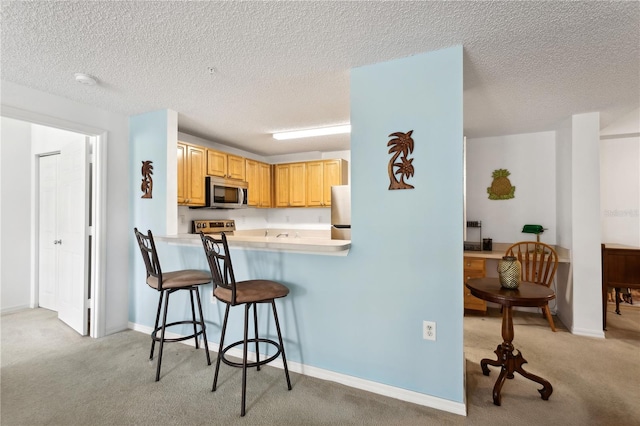 kitchen with light brown cabinetry, a textured ceiling, light colored carpet, a kitchen bar, and appliances with stainless steel finishes