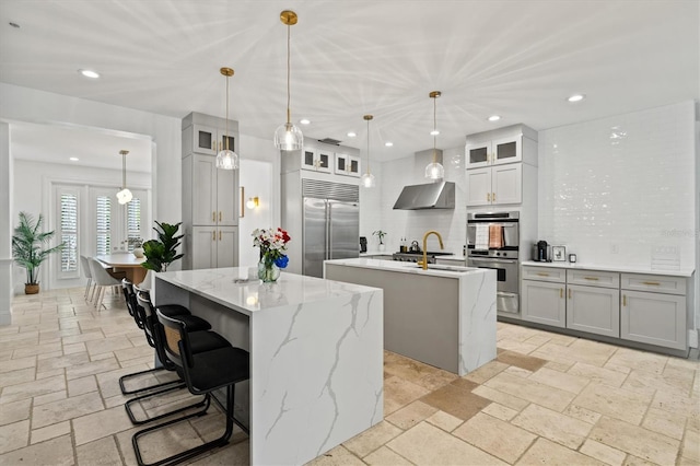 kitchen featuring wall chimney exhaust hood, stainless steel appliances, tasteful backsplash, decorative light fixtures, and a kitchen island with sink