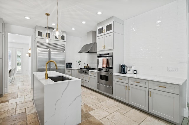 kitchen featuring gray cabinetry, a center island with sink, wall chimney range hood, sink, and appliances with stainless steel finishes