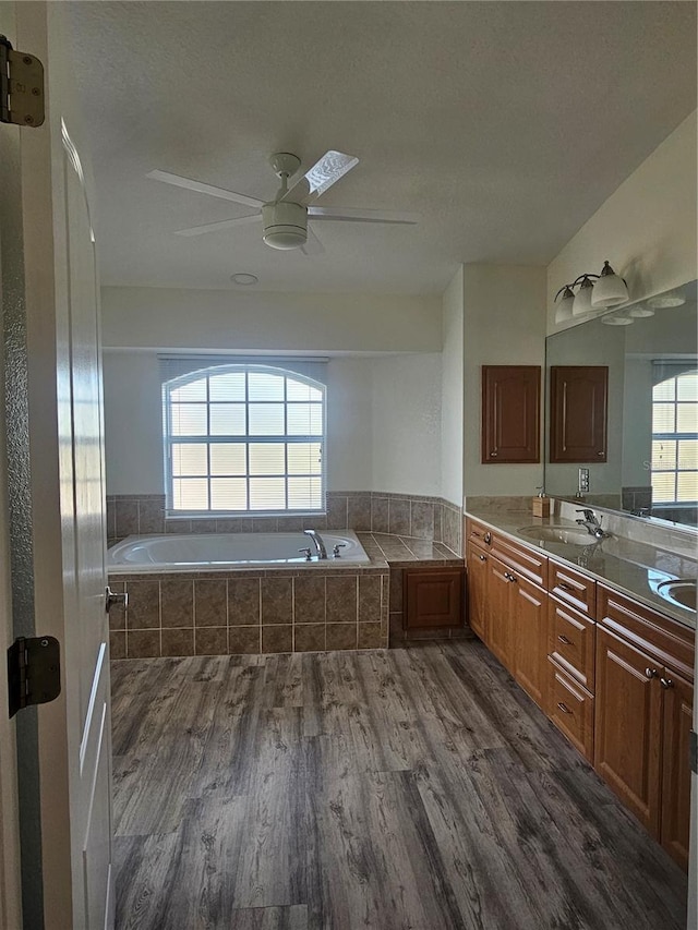 bathroom with ceiling fan, double sink vanity, tiled bath, and wood-type flooring