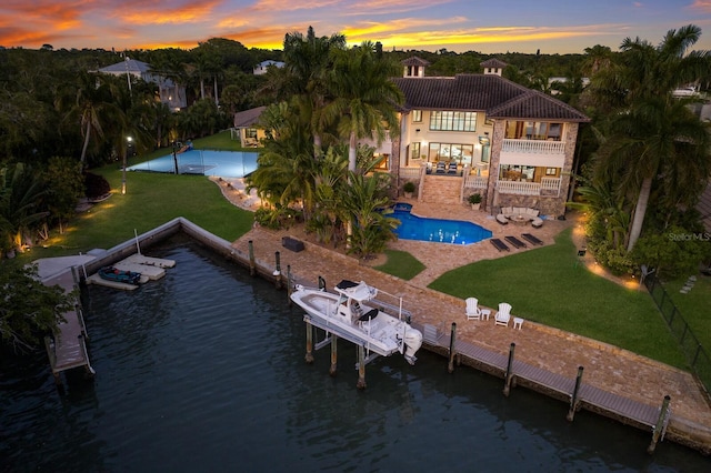 back house at dusk with a water view, a balcony, a patio, and a lawn